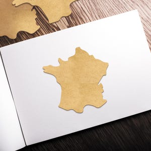 Peut inclure: D&eacute;coupe en bois marron clair en forme de la France, plac&eacute;e sur une surface blanche. La d&eacute;coupe est centr&eacute;e sur la page, avec quelques autres d&eacute;coupes visibles en arri&egrave;re-plan. L'arri&egrave;re-plan est un grain de bois fonc&eacute;.