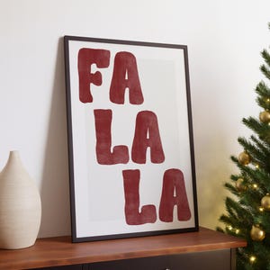 May include: A framed print with the words "FA LA LA" in a bold, red font. The artwork is displayed on a wooden surface, with a decorative vase to the left and a Christmas tree with gold ornaments to the right.