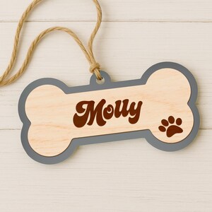 Pode incluir: Uma etiqueta de madeira em forma de osso de c&atilde;o com o nome "Molly" em escrita castanha. A etiqueta tem uma borda cinza e uma pegada de pata. Um cord&atilde;o de barbante natural &eacute; anexado para pendurar.