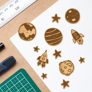 May include: A set of brown wooden cutouts with a space theme. The set includes planets, stars, and a rocket ship. Each cutout has a cute, cartoon-like face.