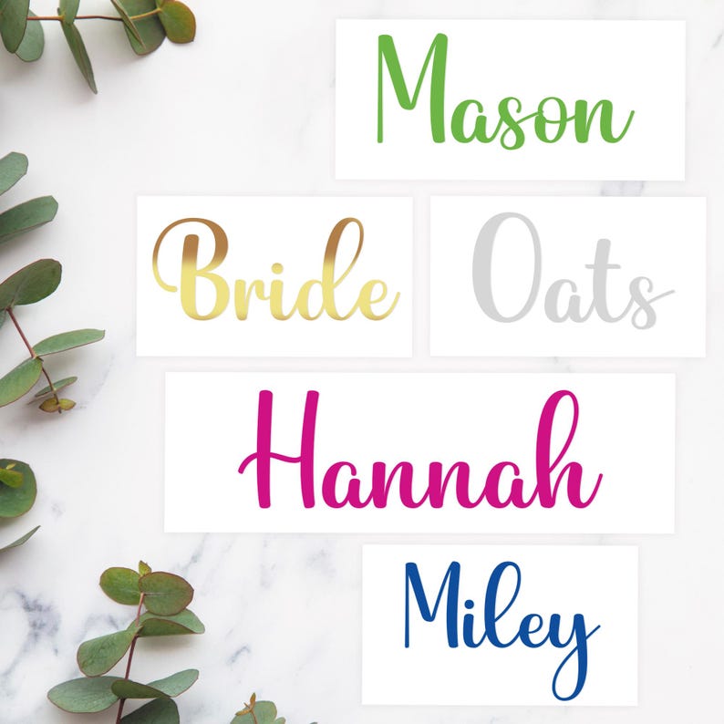 May include: Rectangular white cards with names in various colors: green "Mason", gold "Bride", silver "Oats", magenta "Hannah", and blue "Miley". Eucalyptus leaves are in the background.