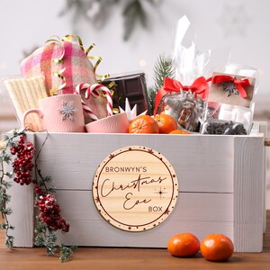 May include: A wooden crate filled with a variety of Christmas Eve treats, including a pink blanket, candy canes, chocolate, and oranges. A round wooden tag with the text "Bronwyn's Christmas Eve Box" is attached to the front of the crate.