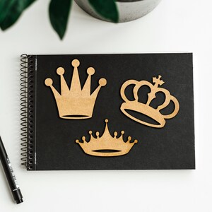 May include: Three wooden crown cutouts in varying designs are arranged on a black spiral-bound notebook. The crowns are a light brown color and have different shapes, including a classic pointed crown and a more ornate design. A black pen is visible in the lower left corner.