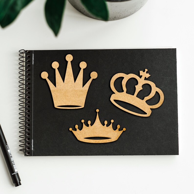 May include: Three wooden crown cutouts in varying designs are arranged on a black spiral-bound notebook. The crowns are a light brown color and have different shapes, including a classic pointed crown and a more ornate design. A black pen is visible in the lower left corner.