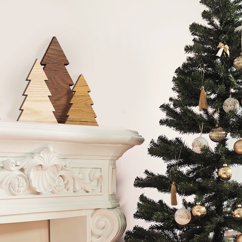 May include: Three wooden Christmas tree ornaments in different sizes and shades of brown and beige, arranged on a white mantelpiece with ornate carvings. A larger green Christmas tree with gold and white ornaments is in the background.