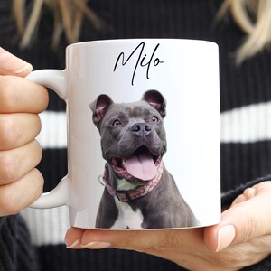 May include: White ceramic mug featuring a gray dog with a pink collar. The dog's name, "Milo," is written in black script above the dog's image. The mug is held by a person.
