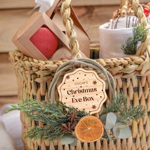 May include: A wicker basket filled with gifts for a Christmas Eve box. The basket is decorated with a wooden tag that says "Oscar's Christmas Eve Box" and a sprig of greenery.