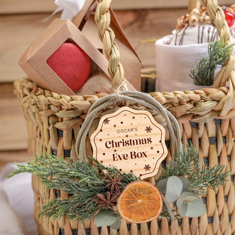 May include: A wicker basket filled with gifts for a Christmas Eve box. The basket is decorated with a wooden tag that says "Oscar's Christmas Eve Box" and a sprig of greenery.