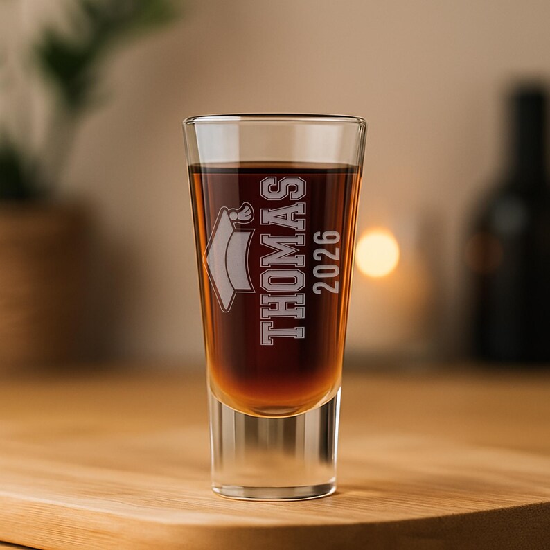 May include: A clear shot glass filled with a dark liquid, possibly liquor. The glass is etched with the name "Thomas" and the year "2026" along with a graduation cap. The glass sits on a wooden surface.