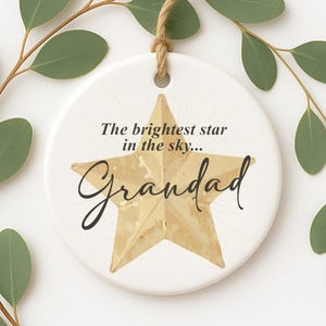 May include: A white ceramic ornament with a gold star design and the words "The brightest star in the sky... Grandad" in black script. The ornament is round with a jute string for hanging.