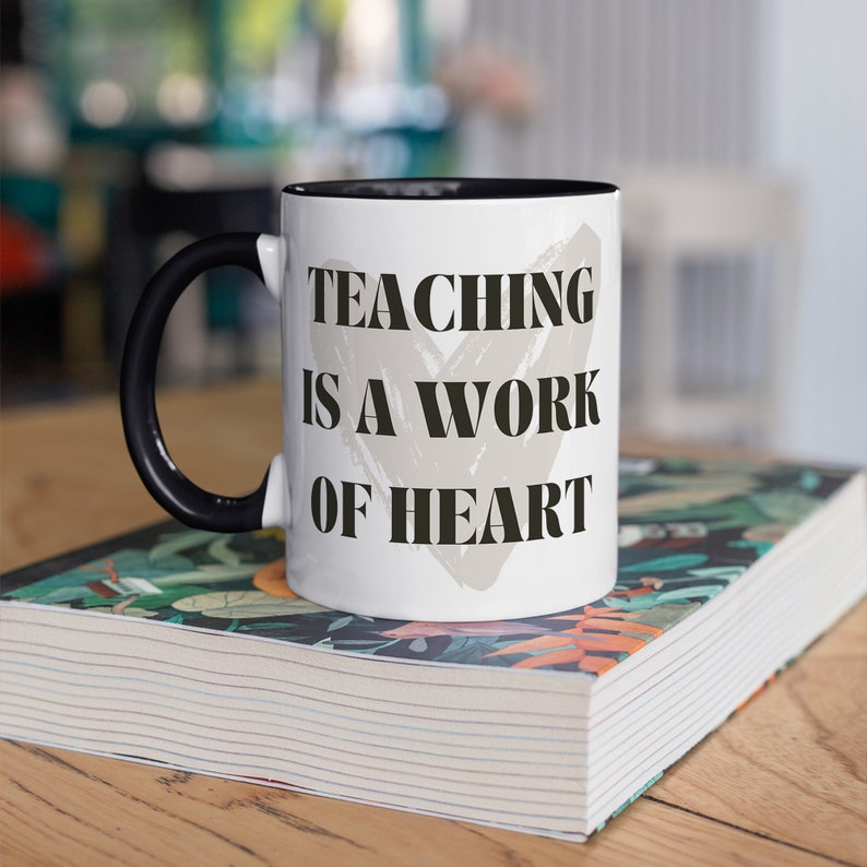 K&ouml;nnte beinhalten: Wei&szlig;e Keramik-Kaffeetasse mit schwarzem Henkel und schwarzem Rand. Die Tasse hat ein graues Herzdesign mit dem Text "TEACHING IS A WORK OF HEART" in Schwarz.