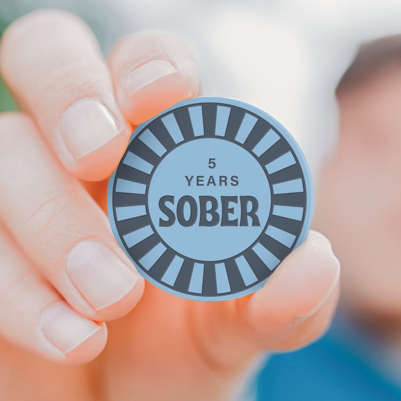 May include: A person is holding a blue and gray round token with the text "5 YEARS SOBER" on it.