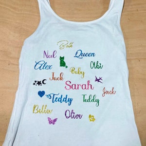 May include: White vest top with multi-coloured glitter text featuring names such as Beth, Jack, and Olive. The design includes a green cat, a heart, a butterfly, and a small airplane.