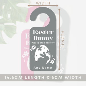 May include: A gray door hanger with a pink border. It features a white bunny silhouette inside an egg with flowers. The text reads "Easter Bunny Please stop here for [Name]" and measures 14.6 cm in length and 6 cm in width.