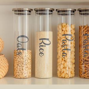 May include: Four clear glass canisters with stainless steel lids, filled with oats, rice, pasta, and penne. The canisters are labeled with the contents in a script font. The oats are at the bottom of the first canister, and the rice is in the second.