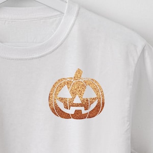 May include: White t-shirt with a gold glitter pumpkin design. The pumpkin has a jack-o'-lantern face.