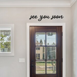 Peut inclure: Une d&eacute;coration murale en m&eacute;tal noir avec l'inscription "see you soon" en &eacute;criture cursive. Le signe est mont&eacute; au-dessus d'une porte en bois fonc&eacute; avec des panneaux de verre. La porte est encastr&eacute;e dans un mur gris clair.