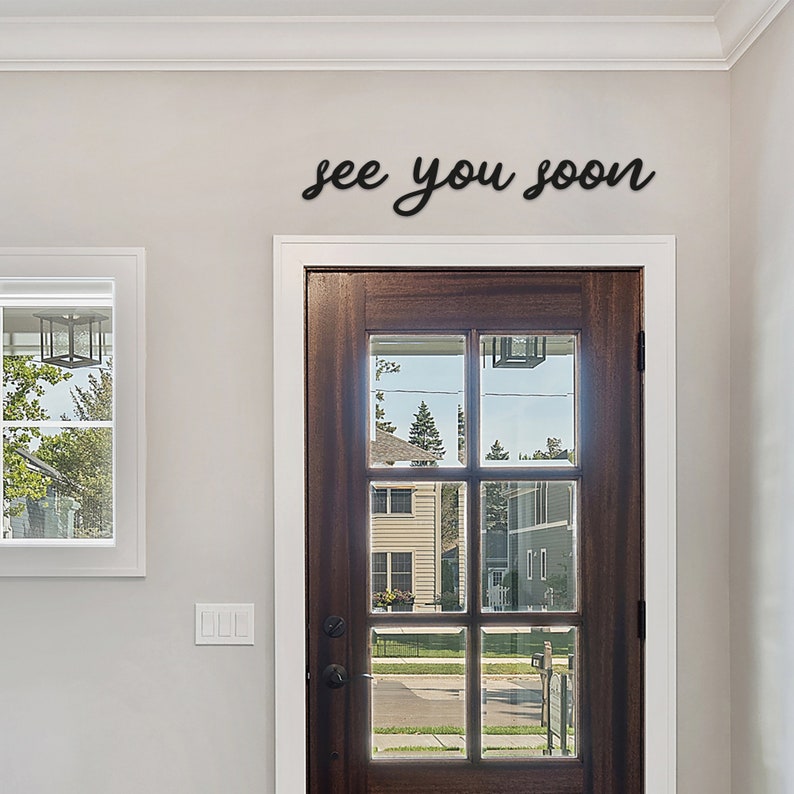 Peut inclure: Une d&eacute;coration murale en m&eacute;tal noir avec l'inscription "see you soon" en &eacute;criture cursive. Le signe est mont&eacute; au-dessus d'une porte en bois fonc&eacute; avec des panneaux de verre. La porte est encastr&eacute;e dans un mur gris clair.