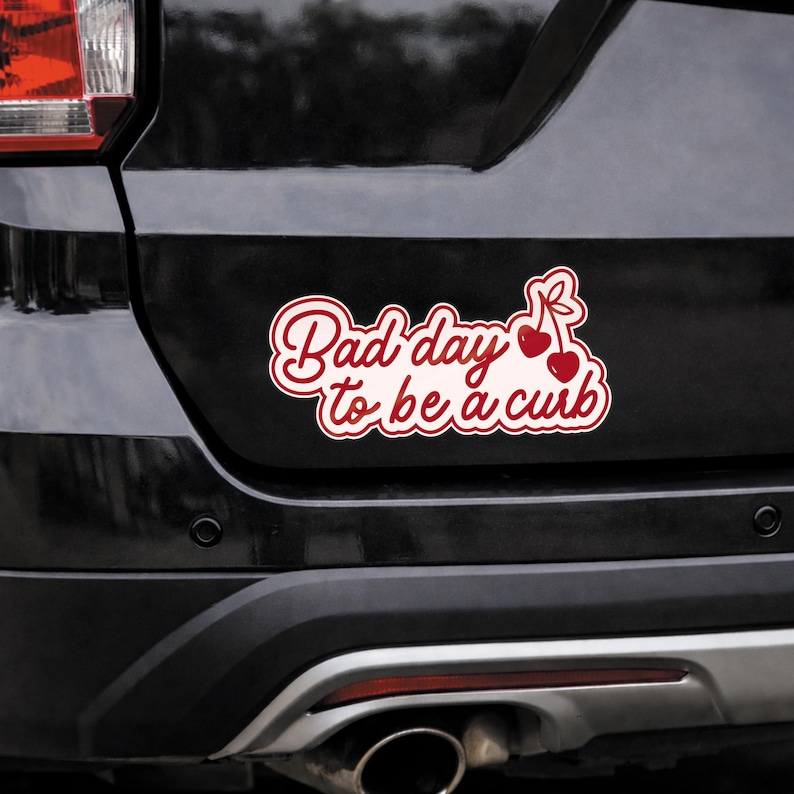 K&ouml;nnte beinhalten: Ein wei&szlig;-roter Autoaufkleber mit dem Text "Bad day to be a curb" in Schreibschrift. Der Aufkleber zeigt auch ein Paar Kirschen mit Stielen. Der Aufkleber ist auf der R&uuml;ckseite eines schwarzen Fahrzeugs angebracht.
