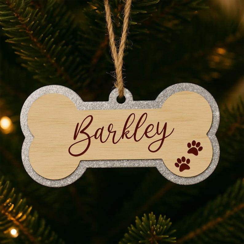 Może przedstawiać: Ozdoba świąteczna w kształcie psiej kości z napisem "Barkley" w bordowym skrypcie. Ozdoba ma jasny, drewniany środek, srebrną obw&oacute;dkę i dwa bordowe odciski łap. Wisi na naturalnym sznurku.