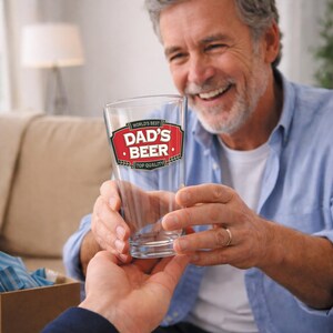 May include: A clear pint glass with the words "WORLD'S BEST DAD'S BEER TOP QUALITY" in a red and white design. The glass is being held by a person. The image is of a gift for Father's Day.