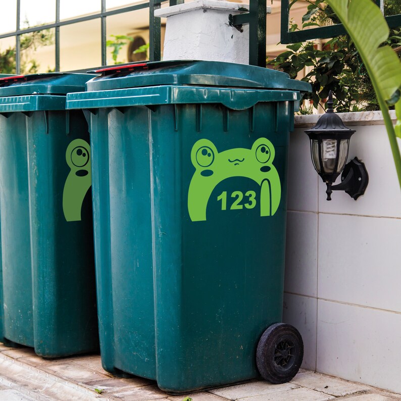 Puede incluir: Dos contenedores de reciclaje verdes con pegatinas de ranas. El contenedor de la derecha tiene el n&uacute;mero 123 dentro de la boca de la rana.