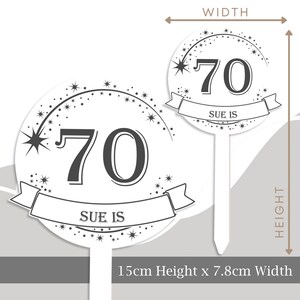 May include: White cake topper with a black and white design. It features the number 70 with a banner that says "Sue is" and a shooting star with a trail of stars. The topper is 7.8 cm wide and 15 cm high.