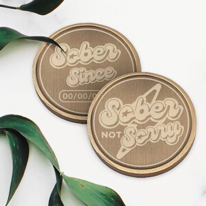 May include: Two gold-colored metal round coins with the words "Sober Since" and "Sober Not Sorry" engraved on them. The coins have a star design and a textured surface.