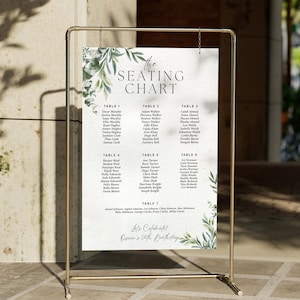 May include: A gold metal frame holds a white seating chart with green botanical accents. The chart lists names under table numbers, with the text "The Seating Chart" at the top and "Let's Celebrate! Oscar's 50th Birthday" at the bottom.
