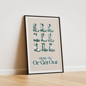 May include: A framed poster with a beige background and a black frame. The poster features a design of nine pairs of cowboy boots with a floral pattern in teal and white. The text "Giddy Up Or Get Out" is printed in teal below the boots.