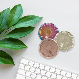 Puede incluir: Tres posavasos redondos con el texto "TEACHING BRIGHT STUDENTS TO SHINE" y un dise&ntilde;o de bombilla. Los posavasos son iridiscentes, oro rosa y dorados. Un teclado blanco y hojas verdes tambi&eacute;n est&aacute;n en la imagen.