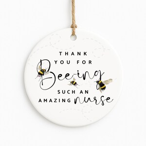 May include: A round, white ceramic ornament with a jute string for hanging. The ornament features the text "Thank You For Being Such An Amazing Nurse" with bee illustrations. The bees are black and yellow.