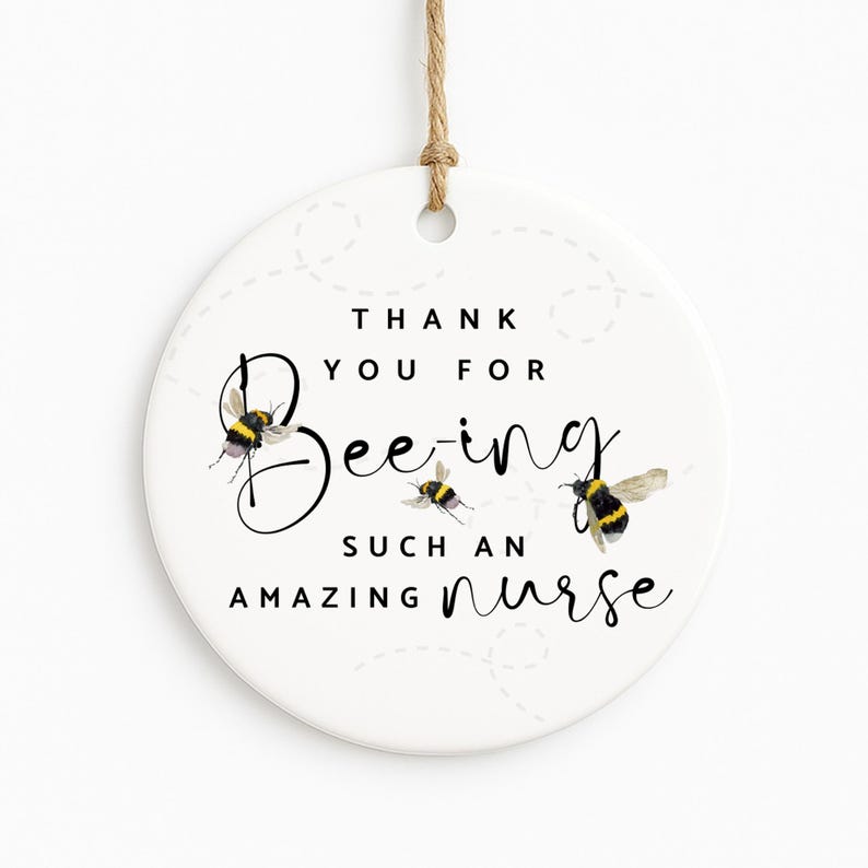 May include: A round, white ceramic ornament with a jute string for hanging. The ornament features the text "Thank You For Being Such An Amazing Nurse" with bee illustrations. The bees are black and yellow.