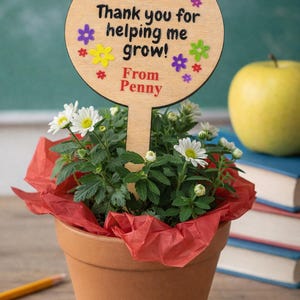 May include: A terracotta pot with a flowering plant and a wooden sign that reads "Thank you for helping me grow! From Penny." The sign is decorated with colorful flower shapes. A yellow apple and books are in the background.