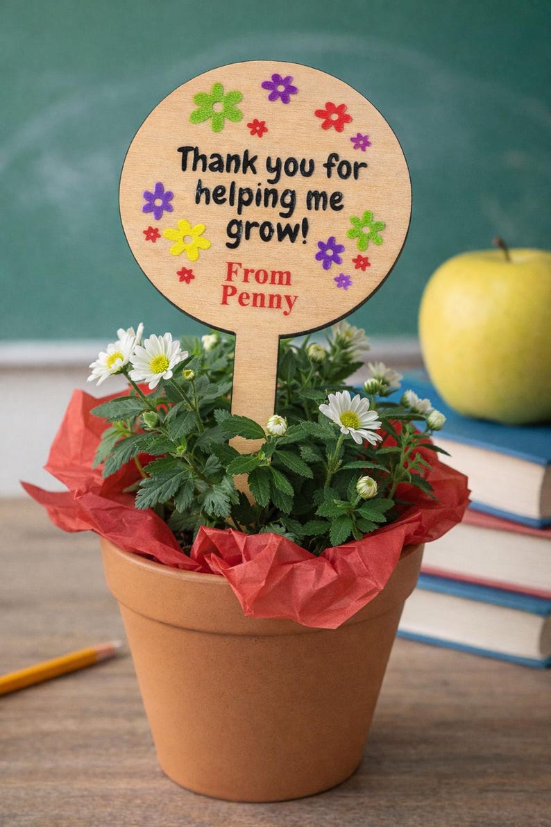 May include: A terracotta pot with a flowering plant and a wooden sign that reads "Thank you for helping me grow! From Penny." The sign is decorated with colorful flower shapes. A yellow apple and books are in the background.