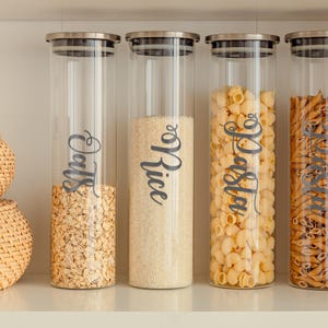 May include: Four glass jars with silver lids filled with various pantry staples. The jars are labeled with black script: "Oats", "Rice", "Pasta", and "Couscous".