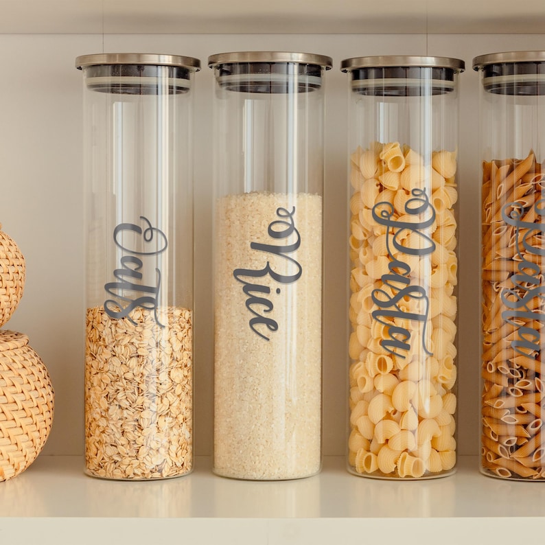 May include: Four glass jars with silver lids filled with various pantry staples. The jars are labeled with black script: "Oats", "Rice", "Pasta", and "Couscous".