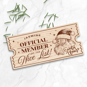 May include: A wooden ticket with a brown wood grain finish. The ticket says "Jasmine Official Member of the Nice List!" with a Santa Claus illustration and the text "Approved by Santa Claus".