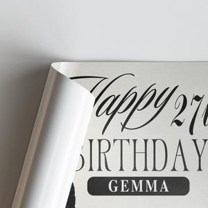 Op de afbeelding: Een lichtgekleurde verjaardagsafdruk met de woorden "Happy 27th Birthday Gemma" in zwart script. De hoek van de afdruk is teruggevouwen en onthult een wit oppervlak.