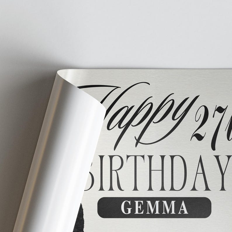 Op de afbeelding: Een lichtgekleurde verjaardagsafdruk met de woorden "Happy 27th Birthday Gemma" in zwart script. De hoek van de afdruk is teruggevouwen en onthult een wit oppervlak.