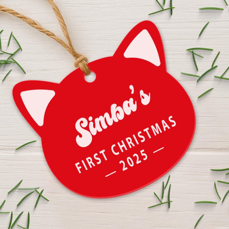 May include: A red cat-shaped Christmas ornament with white accents and a jute string. The ornament reads "Simba's FIRST CHRISTMAS - 2025" in white script. The background includes sprigs of greenery.