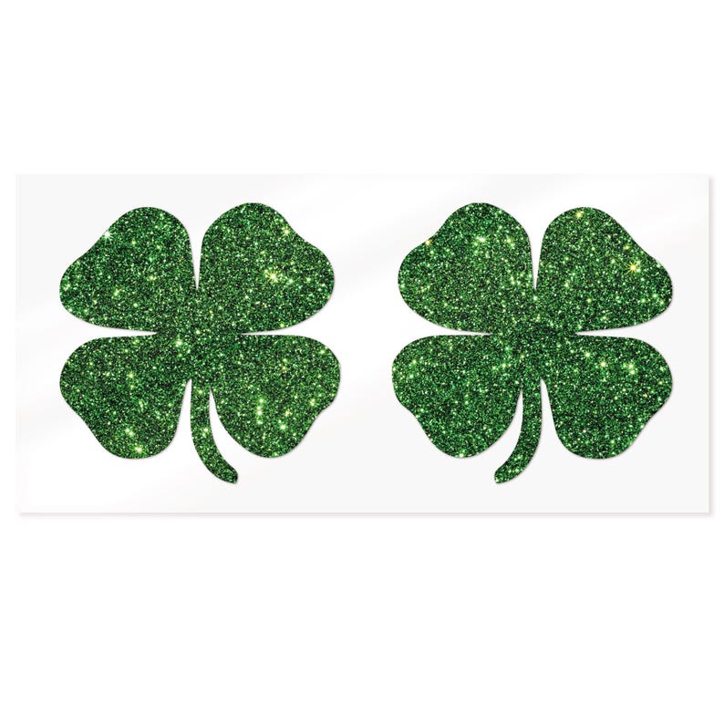 May include: Two green glitter four-leaf clover stickers on a white background. Each clover has four heart-shaped leaves and a stem. The glitter gives a sparkling effect.