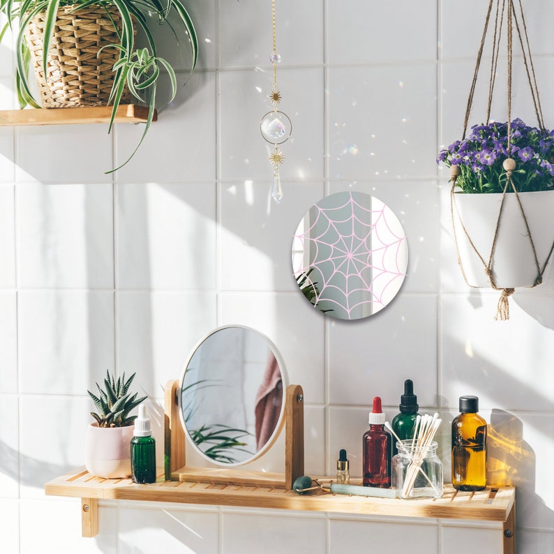 May include: A white bathroom vanity with a wooden shelf, a round mirror with a pink spiderweb design, a smaller round mirror, a potted succulent, and various bottles and jars.