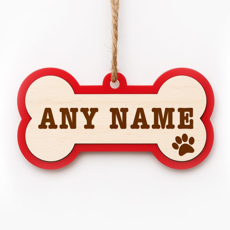 Peut inclure: Ornement en bois en forme d'os de chien, bord&eacute; de rouge et avec un centre en bois clair. Le texte "ANY NAME" est en lettres marron, et une empreinte de patte marron est sur le c&ocirc;t&eacute; droit. Une boucle en ficelle est en haut pour suspendre.