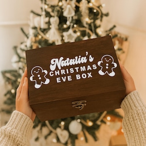 May include: A dark brown wooden Christmas Eve box with the text "Natalia's CHRISTMAS EVE BOX" in white. Two white gingerbread man graphics flank the text. The box has a metal clasp.