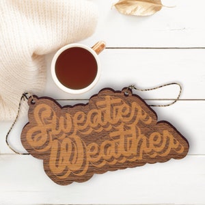 May include: A wooden sign with the words "Sweater Weather" in a decorative font. The sign is brown with lighter brown lettering and a jute string for hanging. A cup of tea and a cream-colored sweater are in the background.