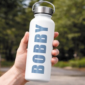 Puede incluir: Una botella de agua blanca con tapa y asa plateadas se sostiene en una mano. La palabra "BOBBY" est&aacute; impresa verticalmente en azul, con letras punteadas. El fondo es follaje verde borroso.