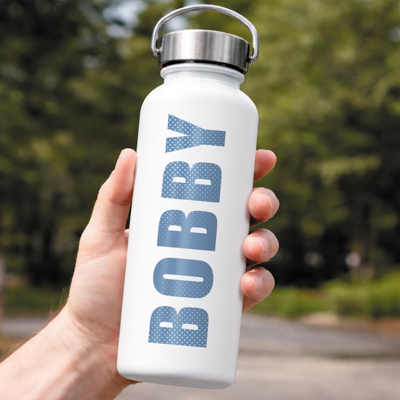 Puede incluir: Una botella de agua blanca con tapa y asa plateadas se sostiene en una mano. La palabra "BOBBY" est&aacute; impresa verticalmente en azul, con letras punteadas. El fondo es follaje verde borroso.