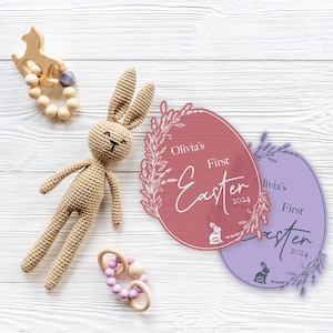 May include: Two pink and purple Easter egg-shaped signs with white floral designs. The signs read "Olivia's First Easter 2024".