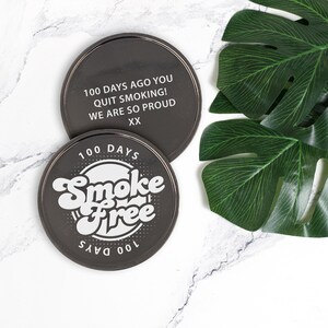 May include: Two black round coasters with white text. The top coaster says "100 days ago you quit smoking! We are so proud XX". The bottom coaster says "100 days Smoke Free 100 days".